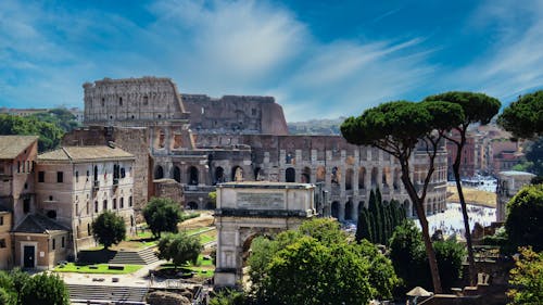 Free From above of aged masonry Colosseum and ancient arch near trees under bright blue cloudy sky in Italy Stock Photo