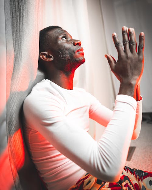 Side view of serious black male in casual outfit sitting on floor near curtain under neon red light and praying while looking up