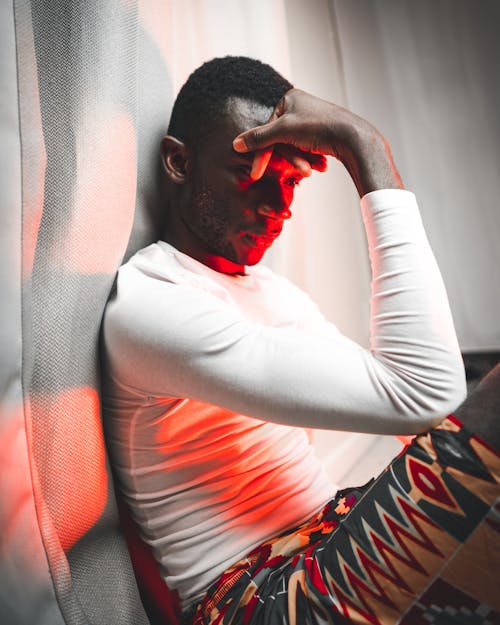 Serious African male in casual outfit sitting near curtain in red neon illumination and looking down pensively