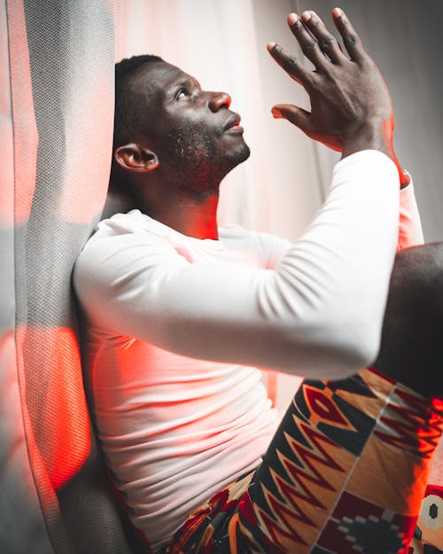 Side view of African male in casual clothes sitting near curtain and praying while looking up thoughtfully