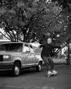 Full length male in casual outfit riding skateboard along road past modern car and trees while recording video on smartphone