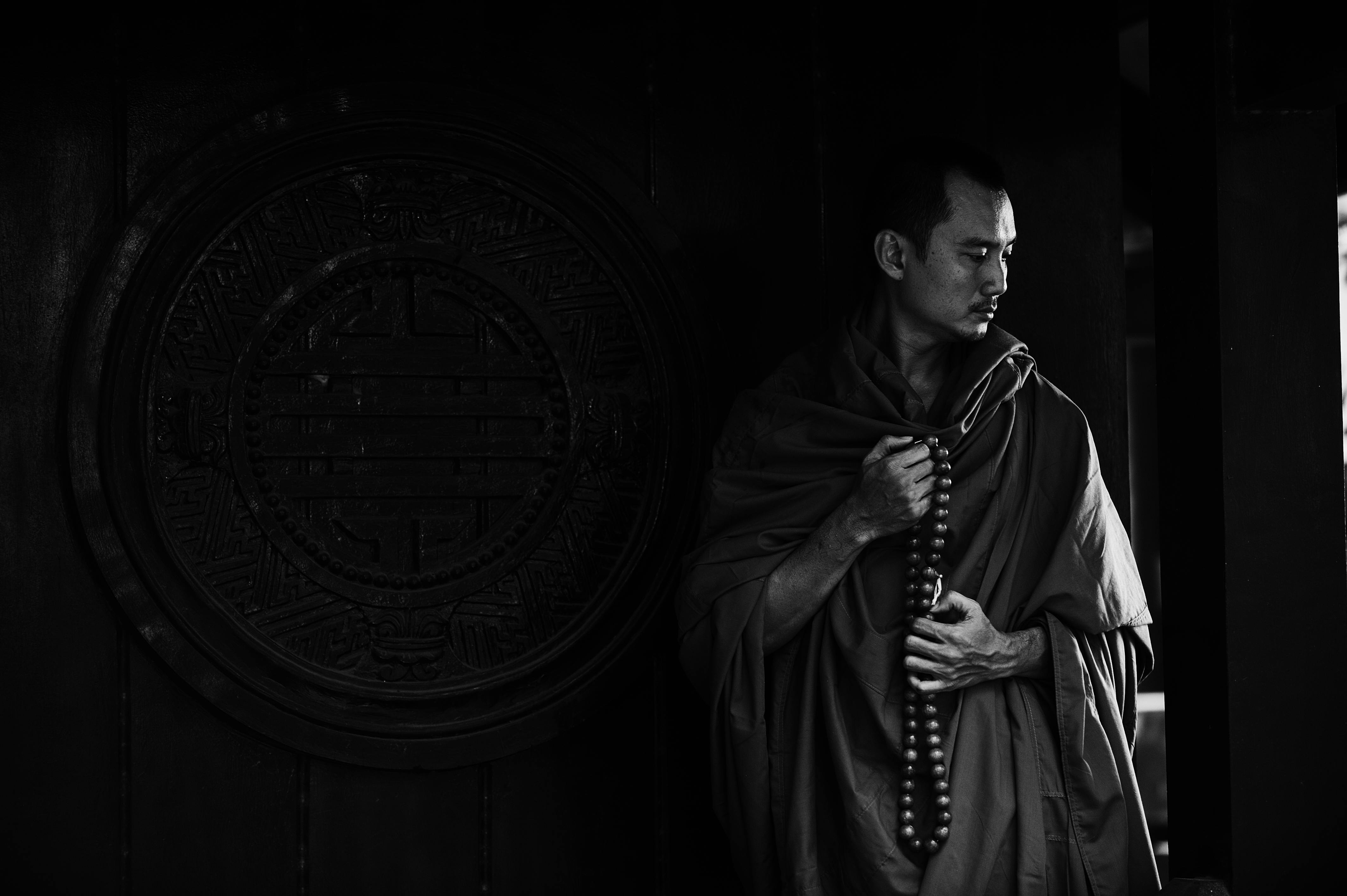 Black and white calm pensive Buddhist monk with beads wearing traditional clothes standing near ornamental temple wall with round caved drawing and looking down in thoughts