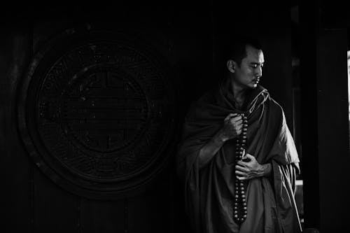 Black and white calm pensive Buddhist monk with beads wearing traditional clothes standing near ornamental temple wall with round caved drawing and looking down in thoughts