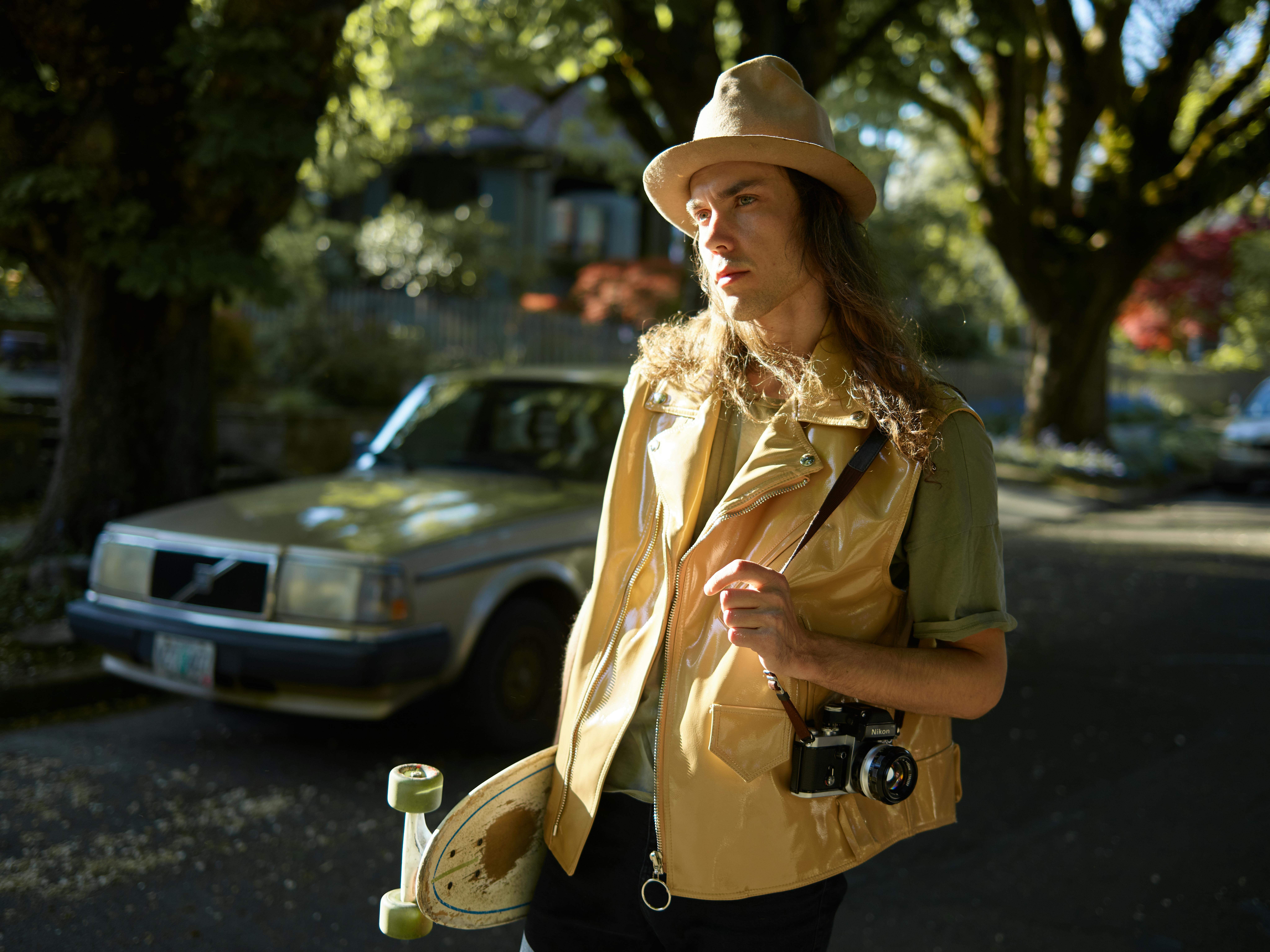Serious long haired male hipster in casual clothes and waistcoat and hat with skateboard in hand and holding camera while standing on street and looking away