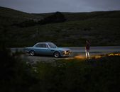 Back view of full length anonymous man in casual outfit standing on roadside in front of parked retro car with lights on near field in twilight