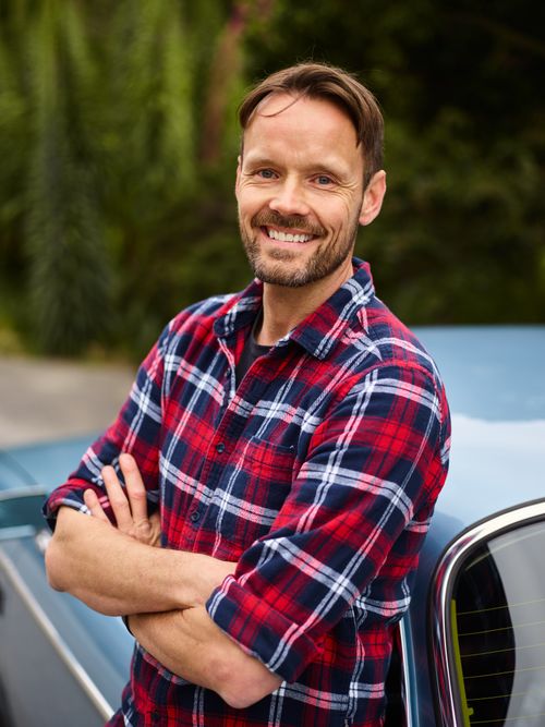 Free Positive middle aged car owner in casual shirt with arms crossed smiling and looking at camera while leaning on automobile against blurred green trees Stock Photo
