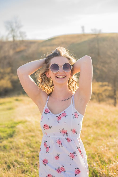 Optimistic smiling female in white summer dress and sunglasses standing with hands behind head on sunny green meadow