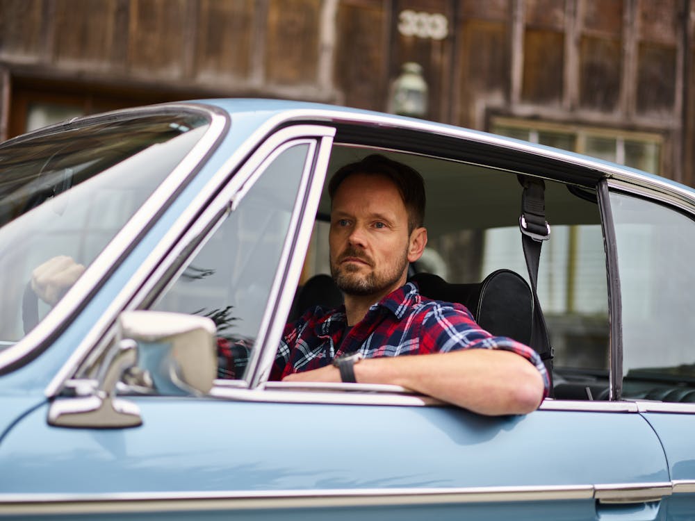 Adult thoughtful bearded driver in wristwatch and casual clothes driving blue automobile while passing by old building and looking away in daylight