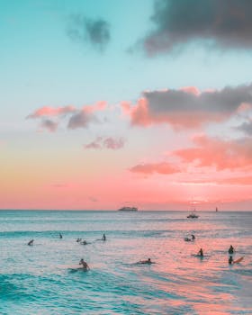 Surfers enjoy the vibrant sunset over Waikiki Beach, with pink skies and ocean waves.