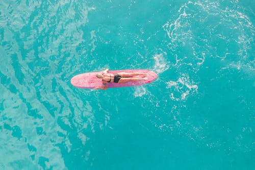 Woman in Pink and White Bikini Lying on Pink Surfboard on Water