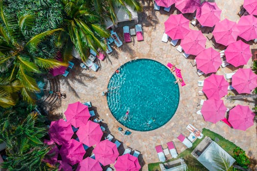 Top-down view of a colorful Waikiki resort pool surrounded by pink umbrellas and lush palm trees.