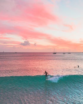 Surfer rides a wave under a vibrant pink sunset in Hawaii. Tranquil ocean scene with sailboats in the distance.