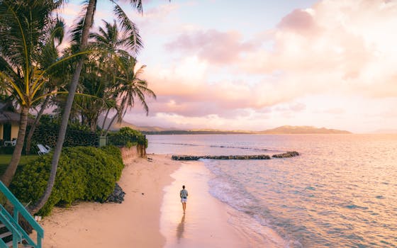 Person walks along Waimanalo Beach, Hawaii at sunrise with palm trees and golden sky.