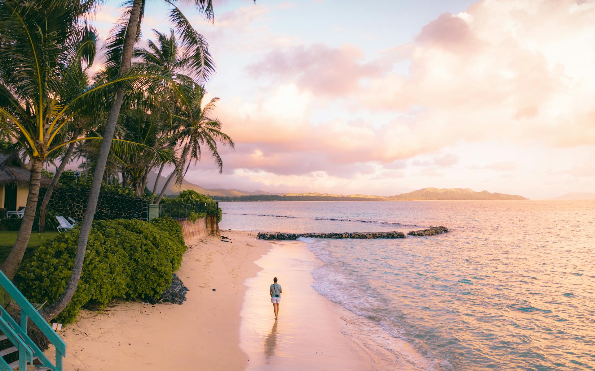 Person walks along Waimanalo Beach, Hawaii at sunrise with palm trees and golden sky.