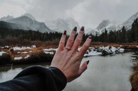 Close-up of a hand wearing an engagement ring with a stunning mountain lake backdrop.