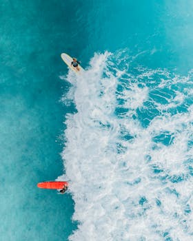 Two surfers catch waves in clear blue waters from an aerial perspective.