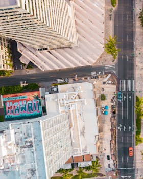 Photo by Jess Loiterton Aerial photograph capturing the vibrant streets of Waikiki, Hawaii with skyscrapers and a 'Greetings from Hawaii' mural.