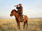 Senior Asian horseman wearing national cloth looking away through vintage monocular standing up in stirrups while riding brown horse in steppe with dried yellow grass and distant mountains in haze ...
