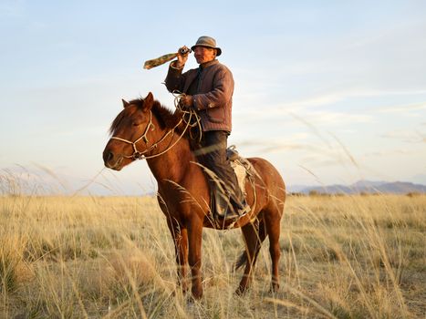 Senior Asian horseman wearing national cloth looking away through vintage monocular standing up in stirrups while riding brown horse in steppe with dried yellow grass and distant mountains in haze under blue sky