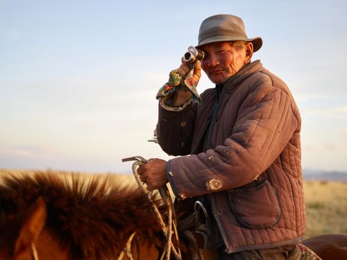 Free Side view of senior Asian male rider wearing padded jacket with holes looking at camera over shoulder through vintage monocular while riding brown horse on blurred background of yellow steppe and blue sky Stock Photo
