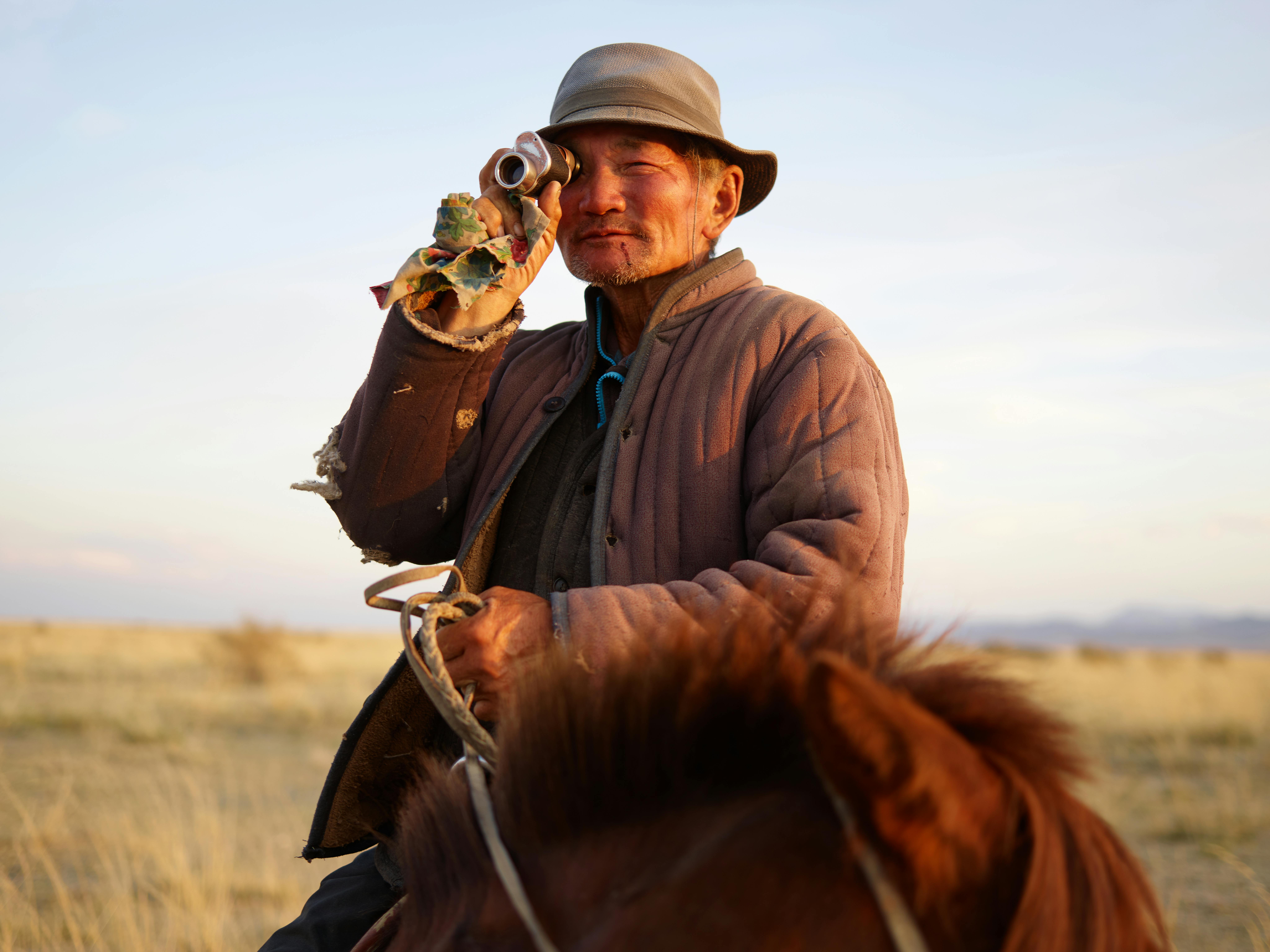 Elderly Asian male farmer wearing old padded jacket looking at camera through vintage monocular while riding brown horse on blurred background of yellow steppe and distant mountains in haze under blue sky