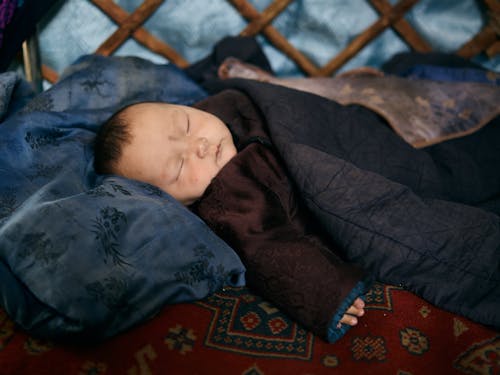 Side view of ethnic baby in warm national clothes sleeping under blanket on place covered by carpet