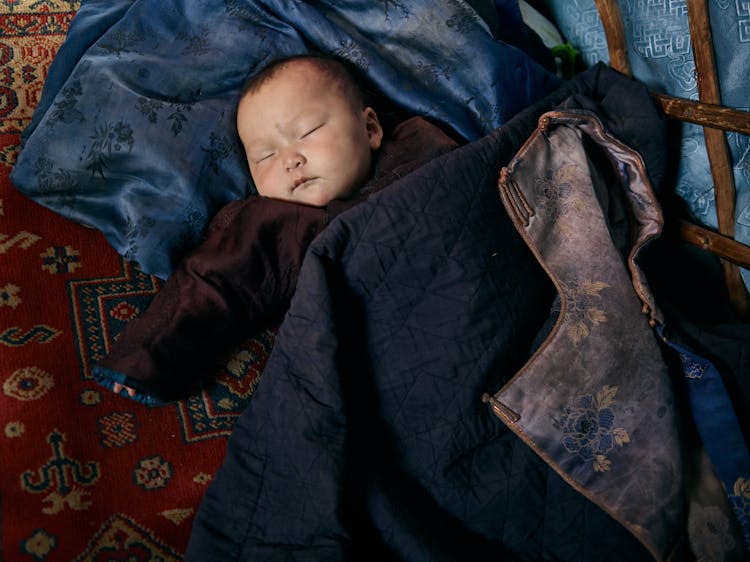 Asian Infant In Warm Clothes Sleeping Under Warm Jacket With Floral Underlay