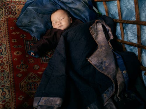 From above of full length sleeping Asian infant in warm clothes lying on carpet under adult man jacket with floral backing