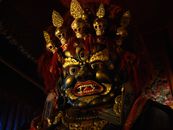 From below of Tsam mystery mask with gilded human face and third eye and pig nose and fangs among teeth and golden circles around face and full head of red hair decorated with red hairband adorned ...