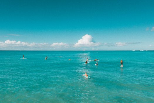 Surfers paddleboarding in the turquoise ocean under a clear blue sky, captured from above.