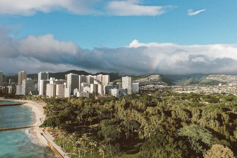 Photo by Jess Loiterton A stunning aerial view of Honolulu with skyscrapers, coastline, and lush greenery under a vibrant sky.