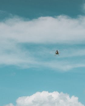 Photo by Jess Loiterton A helicopter flying high against a backdrop of blue sky and white clouds.