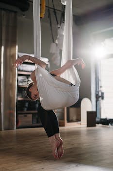 A woman performing an aerial yoga pose, gracefully suspended in a white hammock in a sunlit studio.