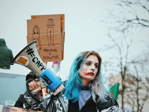 Eccentric young female with blue hair and painted face standing on street with loudspeaker and banner during protest action