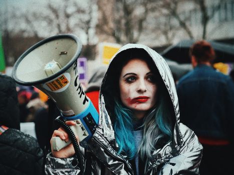 Young informal female in warm clothes with paint on face and blue hair standing on street with loudspeaker in hand among crowd during protest exploration