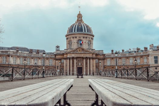 Low angle of famous empty Pont des Arts bridge leading to historic Institut de France building with dome and ornamental walls against cloudy blue sky in Paris