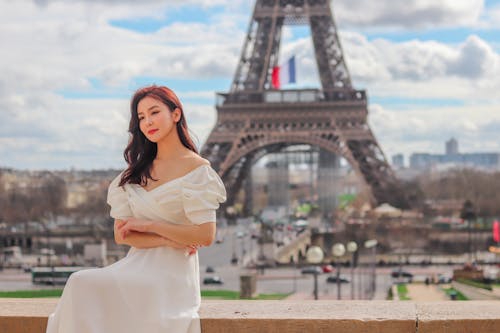 Young romantic Asian female traveler in white dress resting on city square on sunny day with amazing view of Eiffel Tower