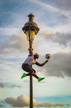 Low angle side view of strong muscular African American male athlete hanging from street old lantern and spinning ball on finger against picturesque cloudy sky