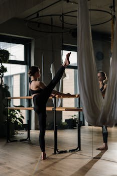 A woman practices barre exercises in a yoga studio with mirrors and hanging fabric.