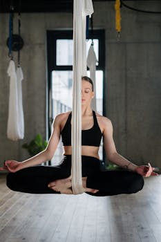 A woman in a yoga studio performing aerial yoga in a suspended lotus pose, focusing on relaxation.