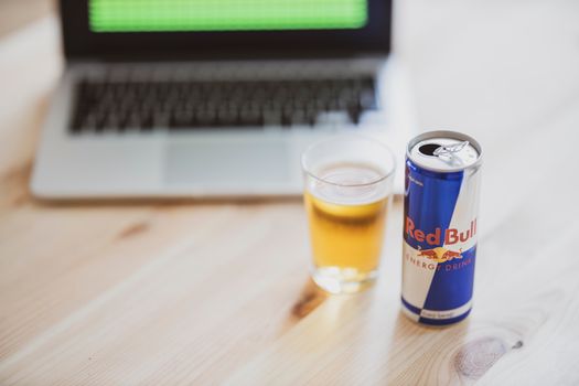 A can of Red Bull and a glass on a wooden desk with a blurred laptop in the background.