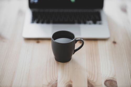 Free Black coffee mug on wooden table with blurred laptop background. Perfect for work-from-home themes. Stock Photo