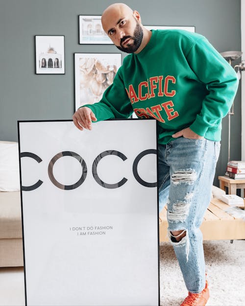 Cool bald male in denim and hoodie holding hand in pocket while standing with framed placard in stylish interior