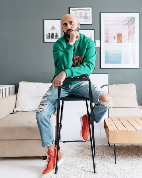 Full length bald male in trendy hoodie and denim leaning on hand while sitting on stool in apartment looking away