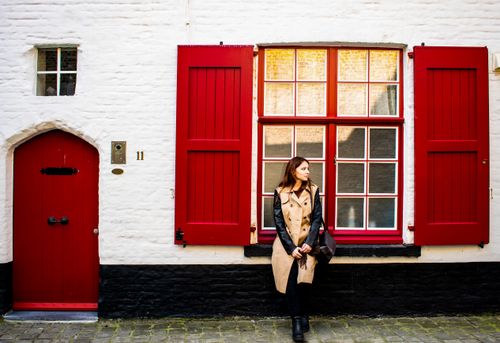 Free Calm pensive female in coat leaning on windowsill with red shutters of building with white brick exterior Stock Photo