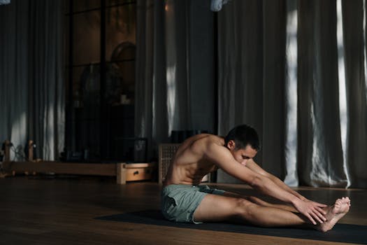 A shirtless man performing seated yoga stretches indoors, focusing on health and wellness.