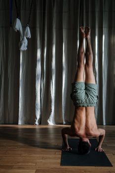 A shirtless man performs a headstand on a yoga mat indoors, demonstrating balance and fitness.