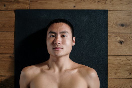 A shirtless man relaxing on a black yoga mat in a wooden floored studio.