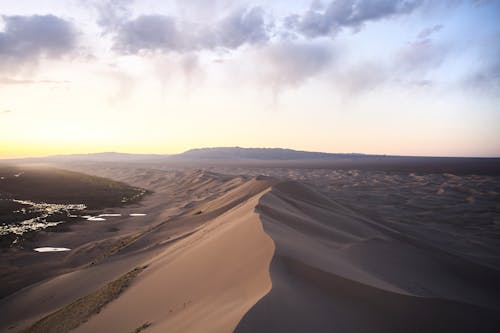 Free Scenery view of sand hills with loose surface located near small puddles and mount behind under colorful sky at sundown Stock Photo