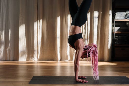 A woman with pink hair performs a handstand on a yoga mat indoors, showcasing strength and balance.